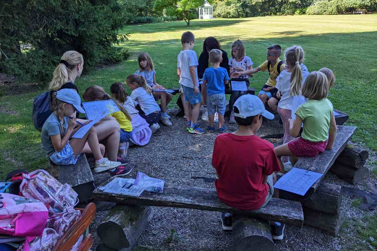 Skupina otrok na jezikovnem poletnem taboru sedi na lesenih klopeh v parku in rešuje delovne liste pod vodstvom mentorice.