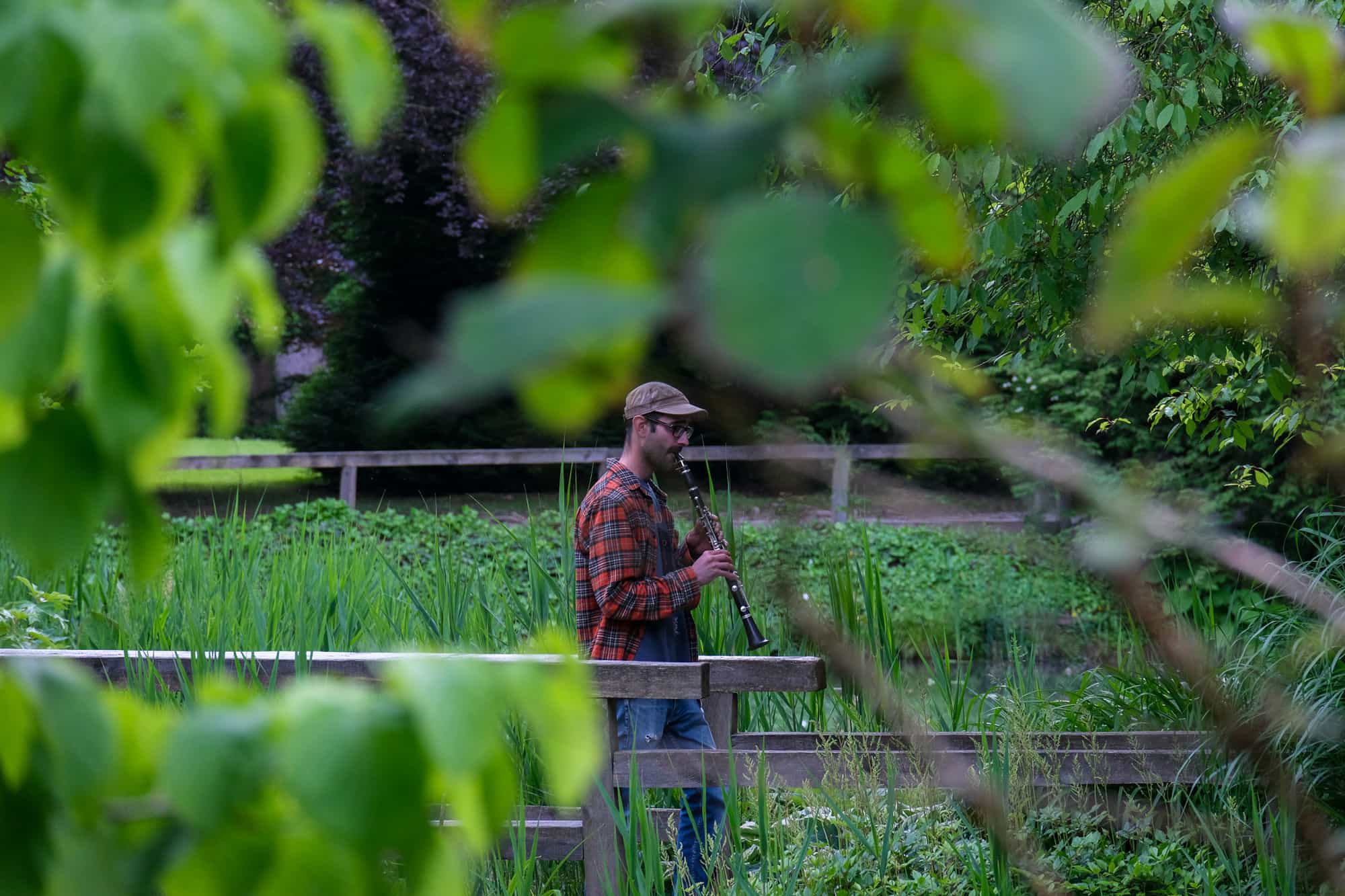 Glasbenik v karirasti srajci igra klarinet na lesenem mostičku sredi gostega zelenega rastlinja in dreves. Pogled na prizor je uokvirjen skozi zamegljene liste v ospredju.