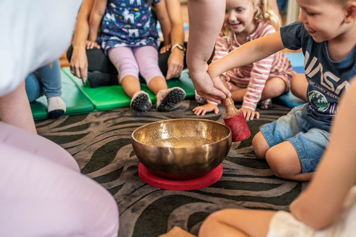 Skupina otrok in odraslih sedi v krogu na vzorčasti preprogi med zvočno terapijo. Odrasla oseba otroku pomaga pri udarjanju po tibetanski zvočni posodi z lesenim tolkalom s kroglo iz rdečega filca.