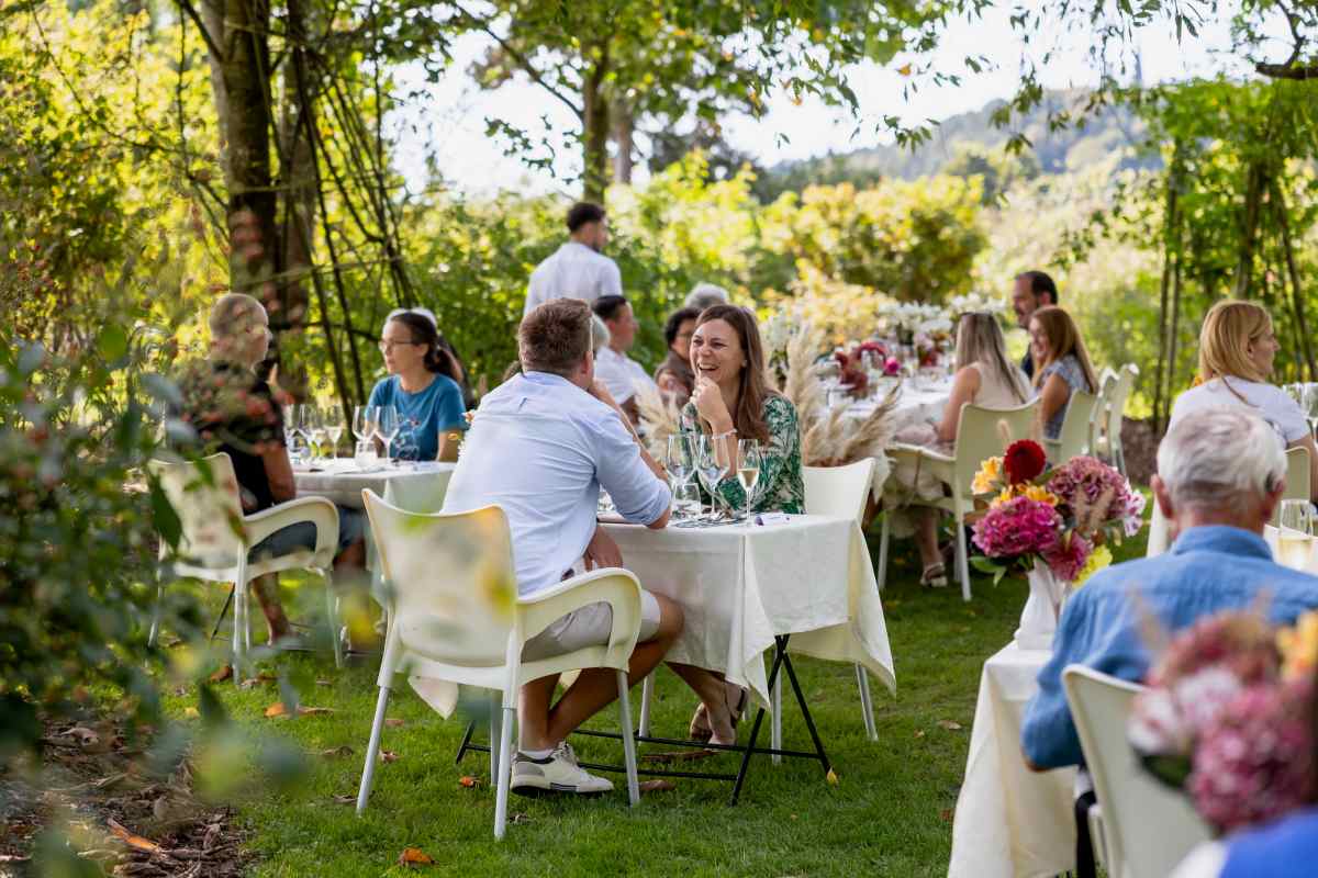 Skupina ljudi uživa ob obedu na prostem v Arboretumu Volčji Potok. Gostje sedijo za belo obloženimi mizami na urejeni trati, obdani z bujnim drevjem in cvetjem. V ospredju se par nasmejan pogovarja.