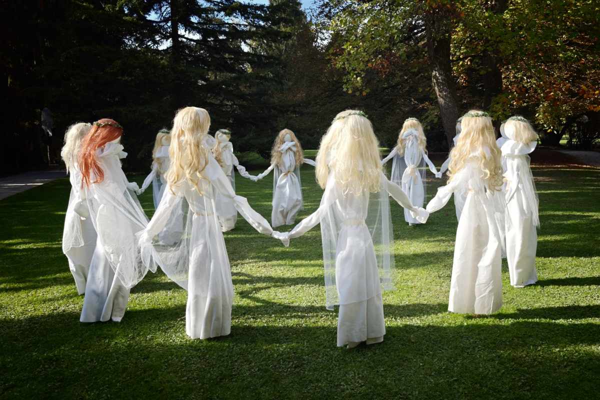 Krog dolgolасih figur v belih oblekah, ki se držijo za roke na trati. Nekatere imajo cvetlične venčke, večina svetle, ena pa rdečkaste lase.
