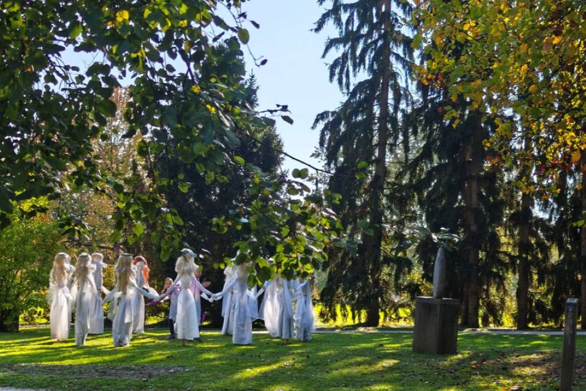 Bele žene pod krošnjami dreves Figure belih žena v dolgih belih oblekah stojijo v krogu na sončni trati pod visokimi drevesi. Med njimi so obiskovalci.