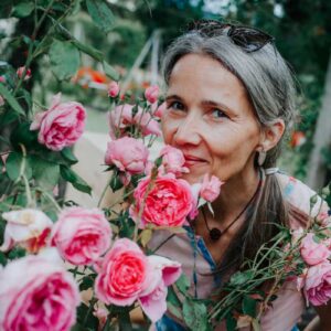 Ženska s spetimi lasmi se sklanja k grmu cvetočih rožnatih vrtnic in jih nežno vonja.