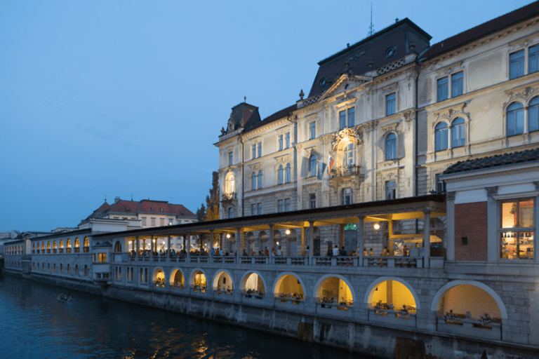Večerni pogled na Plečnikovo tržnico ob Ljubljanici s poudarkom na stavbi Kresije in osvetljenih arkadah, v katerih so ljudje.