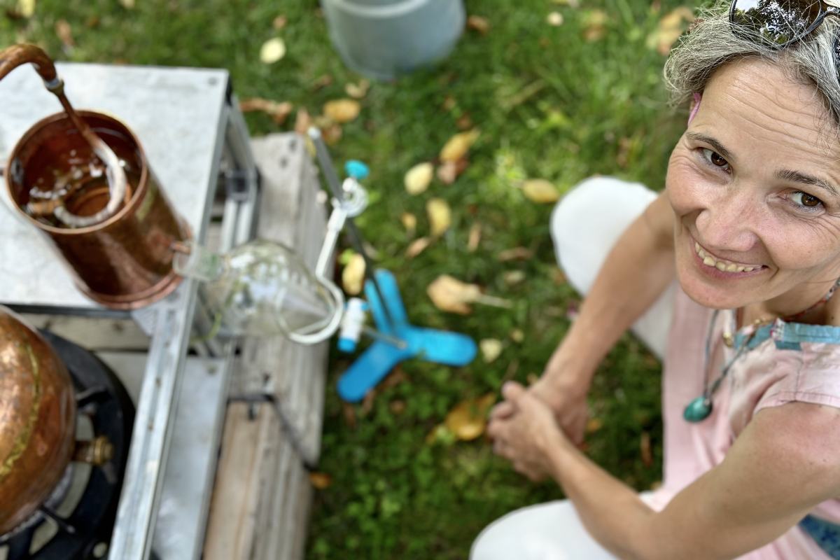 Pogled od zgoraj na nasmejano žensko, ki na travi pripravlja prikaz destilacije rastlin z bakrenim alembikom in stekleno posodo.