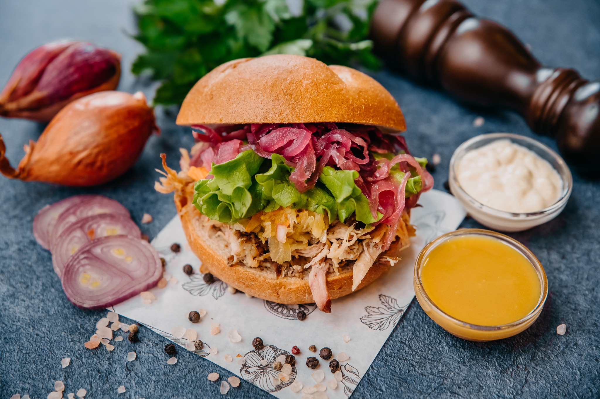 Burgvič s solato, mesom in rdečo čebulo je postrežen na papirju, ob njem pa stojita dve omaki.