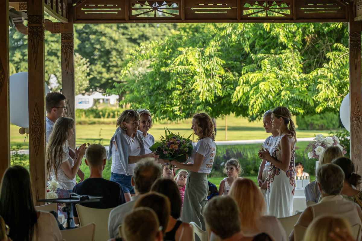 Skupina ljudi v lesenem paviljonu na prostem med podelitvijo šopka rož. V ozadju se razprostira sončen zelen park z drevesi.