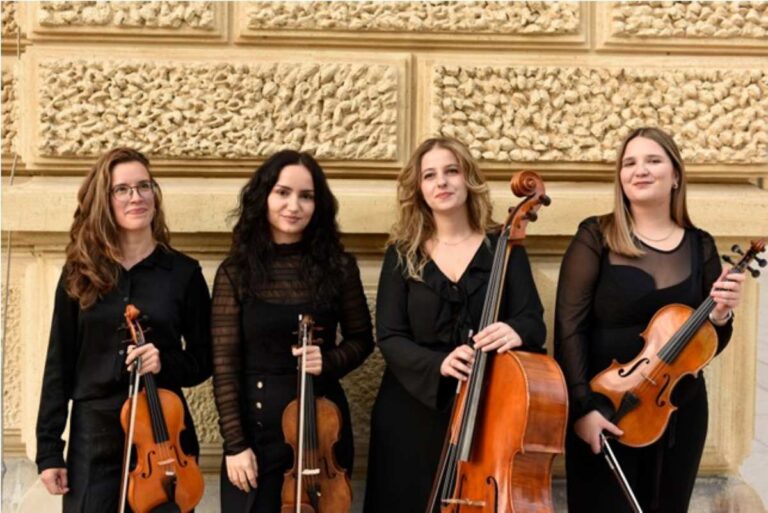 Štiri glasbenice Godalnega kvarteta Apricity v črnih oblačilih pozirajo s svojimi inštrumenti - tremi violinami in violončelom, pred kamnitim zidom z reliefno teksturo.