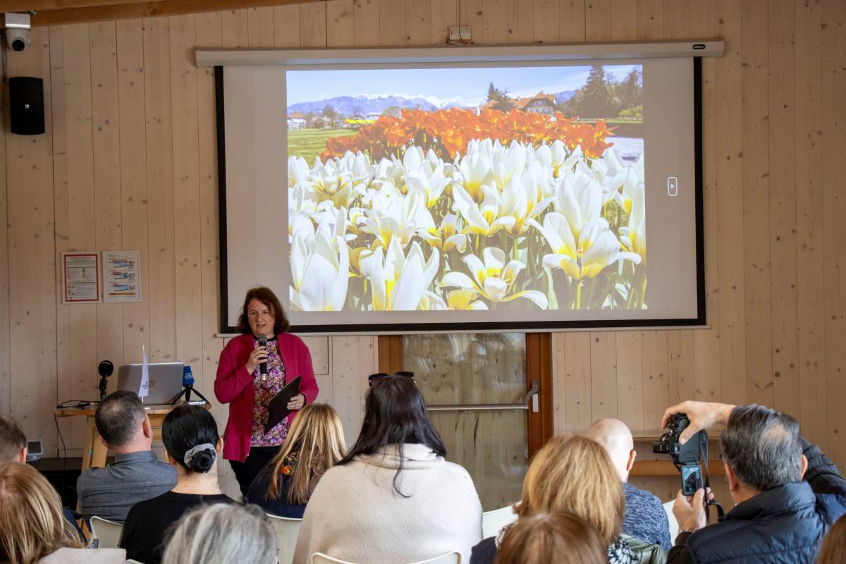 Mateja Račevski na novinarski konferenci v Arboretumu : Mateja Račevski nagovarja občinstvo na novinarski konferenci v Arboretumu Volčji Potok. Za njo je na projekcijskem platnu prikazana fotografija cvetočih tulipanov, obiskovalci pa dogodek spremljajo iz dvorane.