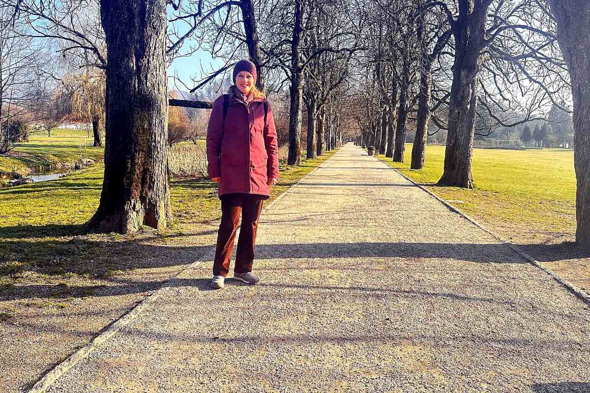 Vesna Resnik v drevoredu Vesna Resnik med obiskom Arboretuma Volčji Potok stoji na dolgem drevoredu, ki ga obdajajo mogočna drevesa in odprti pogledi na park. Fotografija poudari urejenost prostora, mirno vzdušje in lepoto sprehoda skozi arboretum.