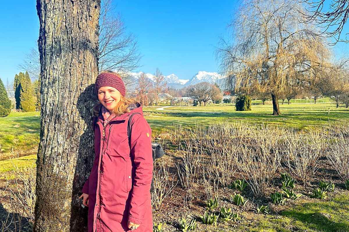 Vesna Resnik med obiskom Arboretuma Volčji Potok stoji ob mogočnem drevesu in se nasmehne v sončen dan. Fotografija povezuje parkovno krajino, drevesa in razgled na zasnežene vrhove v ozadju.