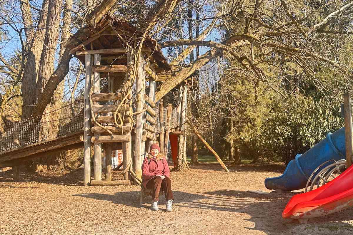 Vesna Resnik na otroškem igrišču Vesna Resnik med obiskom Arboretuma Volčji Potok sedi na otroškem igrišču ob velikih lesenih igralih. Fotografija prikazuje igriv del parka, kjer se naravno okolje prepleta s prostorom za družinsko doživetje in sproščen obisk.