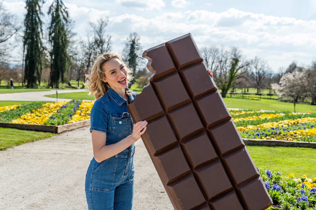 Nasmejana ženska stoji na poti v parku in drži zelo veliko tablico čokolade, okoli nje pa cvetijo rumene in modre pomladne gredice.