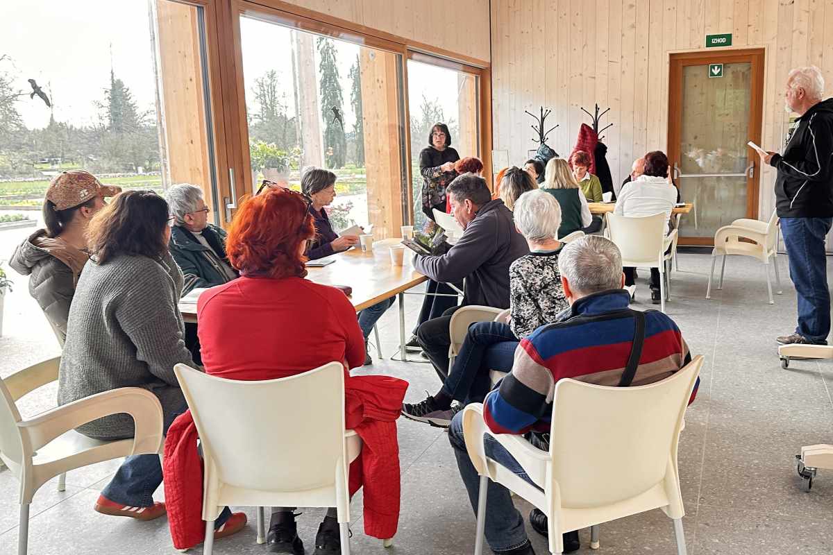 Mija Jerman o pomenu fotografije na usposabljanju Na fotografiji Mija Jerman udeležencem predstavlja pomen fotografije v okviru tretjega usposabljanja za spremljevalce na Poti za spomin v Arboretumu Volčji Potok. Udeleženci sedijo za mizami v svetlem prostoru in spremljajo njeno razlago.