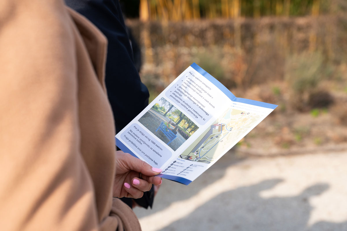 Zloženka Poti za spomin v Arboretumu Volčji Potok Obiskovalka v roki drži zloženko Poti za spomin z zemljevidom in informacijami o poti v Arboretumu Volčji Potok.