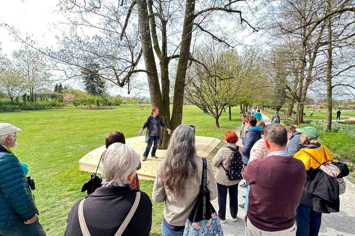 Predstavitev Poti za spomin ob drevesih Skupina obiskovalcev stoji ob poti v Arboretumu Volčji Potok in posluša vodnico, ki stoji na leseni ploščadi okoli velikega drevesa sredi odprte travnate parkovne površine.