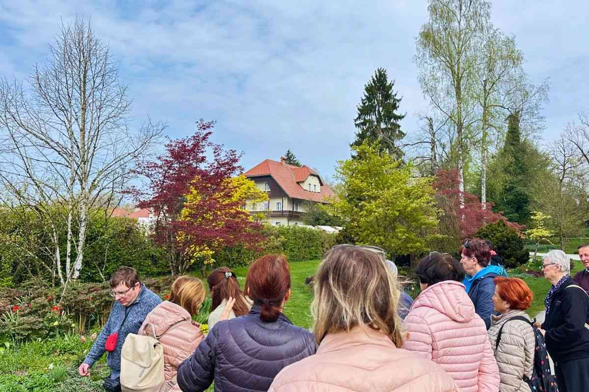 Razgledi in odprtost krajine v Arboretumu Skupina ljudi stoji ob travniku in okrasnih drevesih ter opazuje odprt pogled proti hiši in spomladansko obarvani parkovni krajini.
