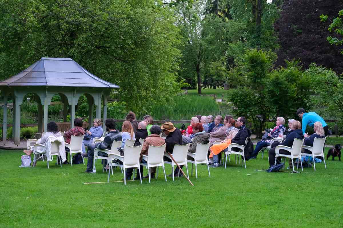 Obiskovalci sedijo na belih stolih na travniku ob paviljonu in prisluhnejo dogodku AnimotMUZIK v zelenem ambientu Arboretuma Volčji Potok.
