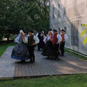 Folklorna skupina v tradicionalnih slovenskih narodnih nošah pleše na lesenem odru na prostem ob stavbi, obdani z zelenjem.