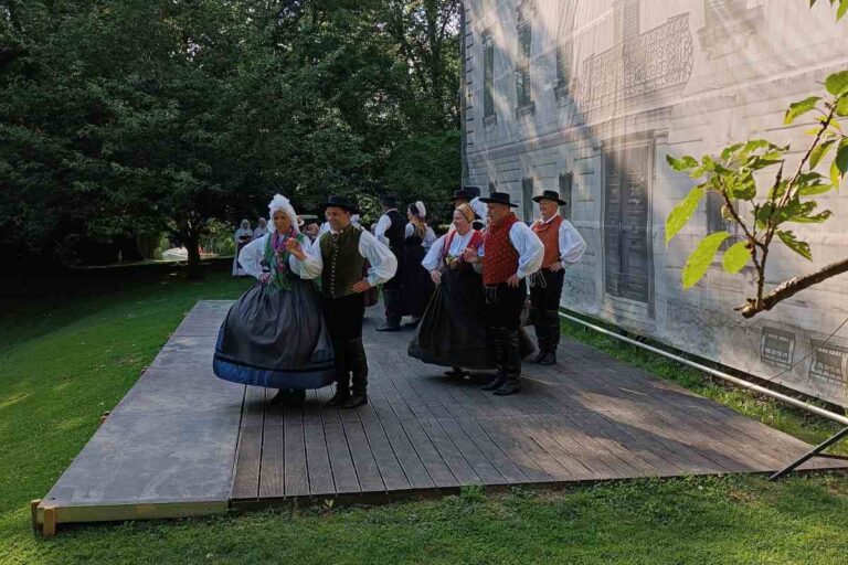 Folklorna skupina v tradicionalnih slovenskih narodnih nošah pleše na lesenem odru na prostem ob stavbi, obdani z zelenjem.