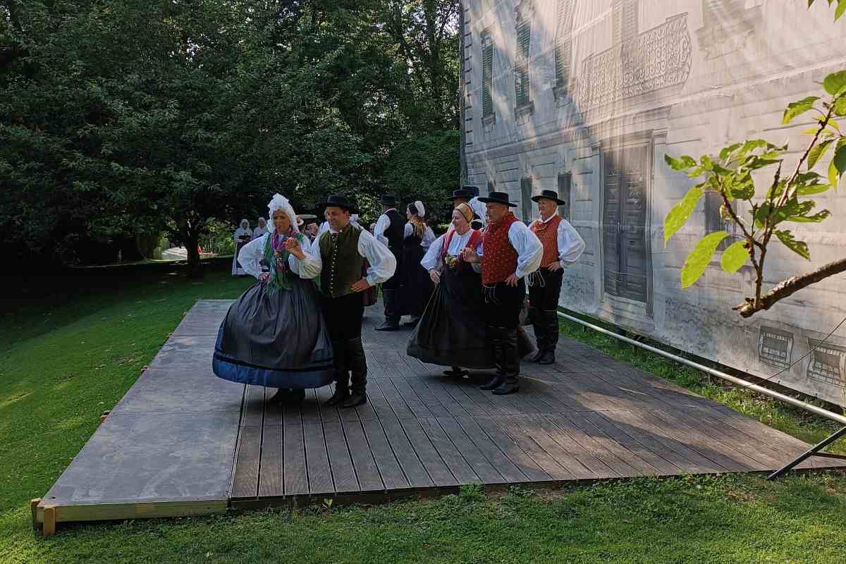 Folklorna skupina v tradicionalnih slovenskih narodnih nošah pleše na lesenem odru na prostem ob stavbi, obdani z zelenjem.