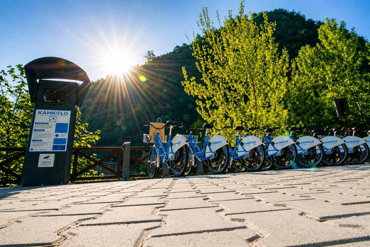 Postaja Kamkolo z vrsto parkiranih mestnih koles pri Arboretumu Volčji Potok v jutranji svetlobi, v ozadju drevesa in sončni žarki.