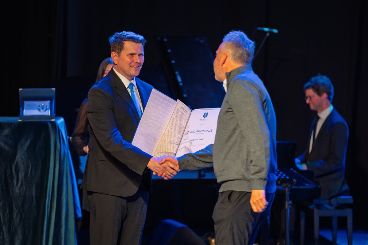 Aleš Ocepek stoji za govorniškim pultom na osvetljenem odru in v rokah drži odprto mapo s priznanjem. Ob pultu je velik šopek belih vrtnic, v ozadju pa stoji evropska zastava.