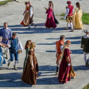 Skupina ljudi v pisanih zgodovinskih kostumih pleše v krogu na prostem. Oblečeni so v oblačila, značilna za renesančno obdobje, in izvajajo usklajene plesne korake v parih in skupinah, kar ustvarja živahno in avtentično vzdušje preteklosti.