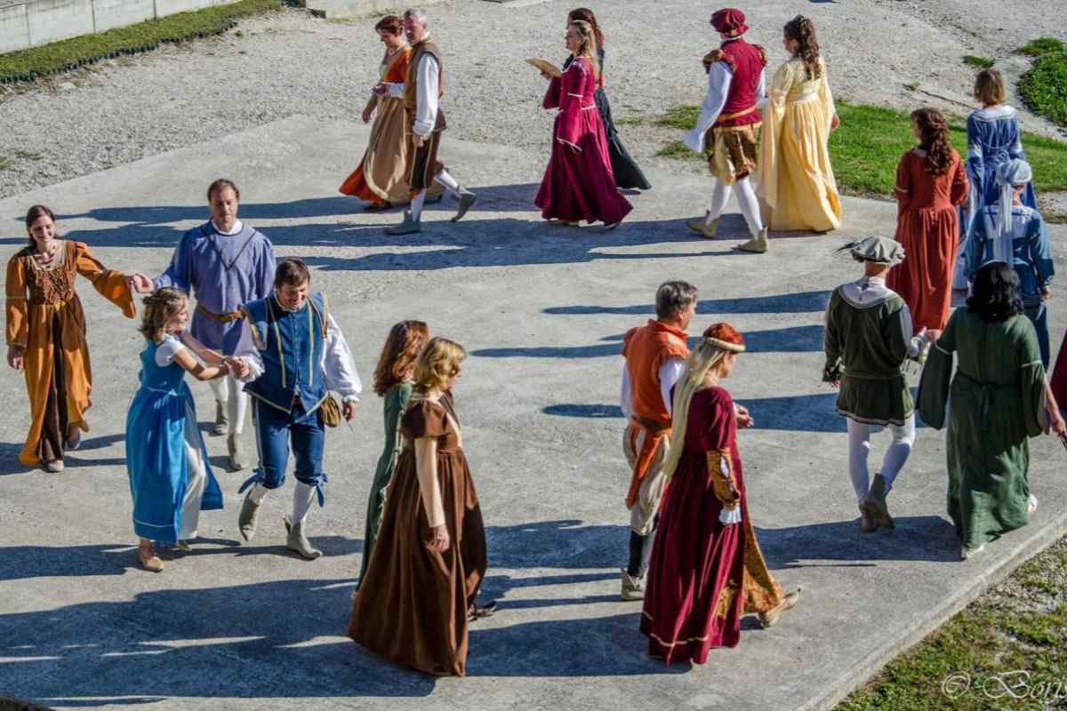 Skupina ljudi v pisanih zgodovinskih kostumih pleše v krogu na prostem. Oblečeni so v oblačila, značilna za renesančno obdobje, in izvajajo usklajene plesne korake v parih in skupinah, kar ustvarja živahno in avtentično vzdušje preteklosti.