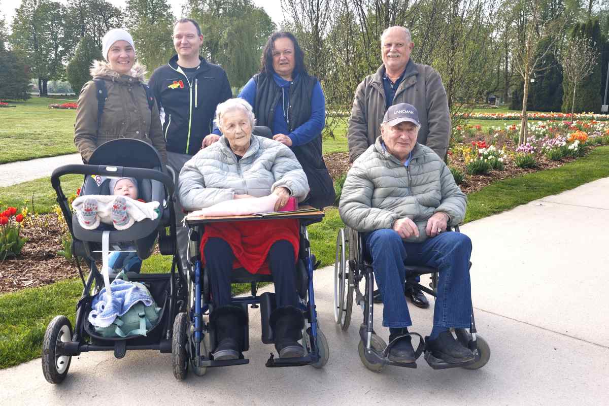 Štiri generacije družine v Arboretumu Volčji Potok Štiri generacije družine stojijo in sedijo na sprehajalni poti v Arboretumu Volčji Potok. V ospredju sta dojenček v vozičku ter starejša ženska in starejši moški na invalidskih vozičkih, v ozadju pa stojijo njuni družinski člani. Ob poti cvetijo tulipani, okoli njih pa se razprostira zelen park.