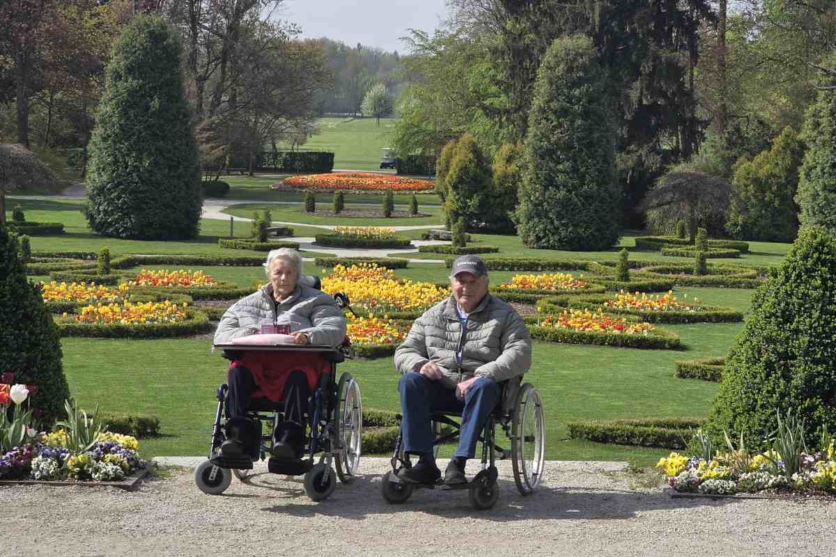 Starejša ženska in starejši moški na invalidskih vozičkih sta fotografirana pred simetrično urejenim Francoskim parkom v Arboretumu Volčji Potok. Okoli njiju so cvetoče gredice, nizko oblikovane žive meje in visoka drevesa.