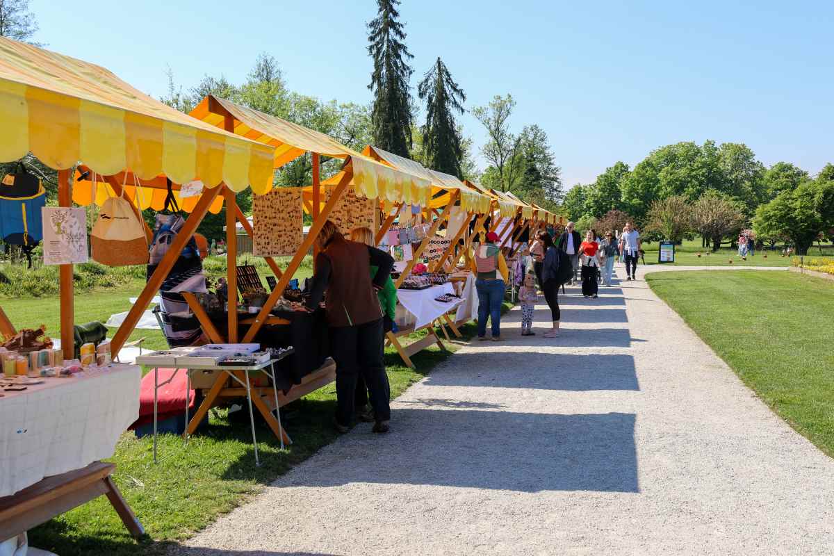 Sejem domače in umetnostne obrti med Spomladansko razstavo v Arboretumu Ob sprehajalni poti v Arboretumu Volčji Potok je postavljena vrsta lesenih stojnic z rumeno belimi nadstreški, ob katerih obiskovalci na sončen pomladni dan obiskujejo sejem domače in umetnostne obrti.