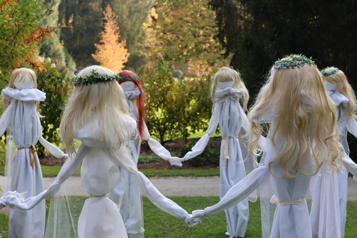 Krog belih žena v jesenskem Arboretumu Figure belih žena v dolgih belih oblekah se držijo za roke v krogu. Imajo dolge lase, nekatere tudi zelene venčke.