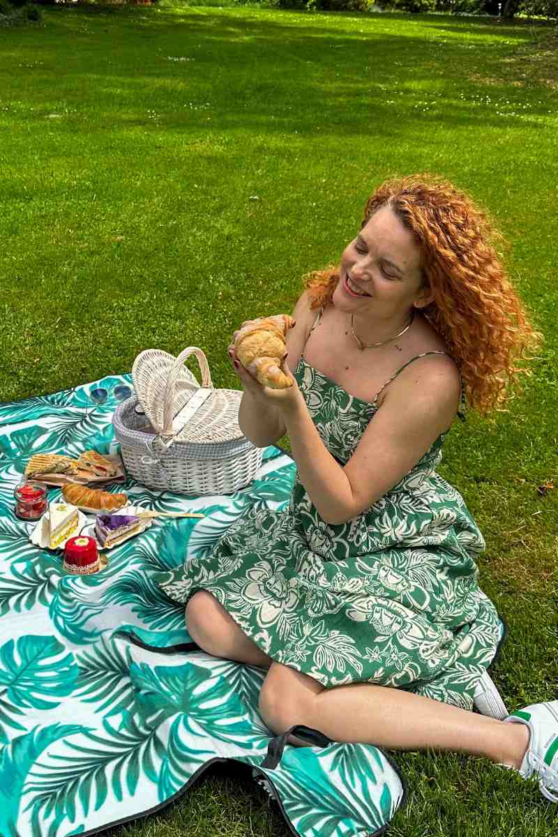 Poletni piknik OBRAZI VRTNIC v parku Nasmejana ženska v zeleni obleki sedi na piknik odeji na travniku. V rokah drži rogljiček, pred njo pa so bela piknik košara, sladice, sendviči in druge dobrote piknik paketa.