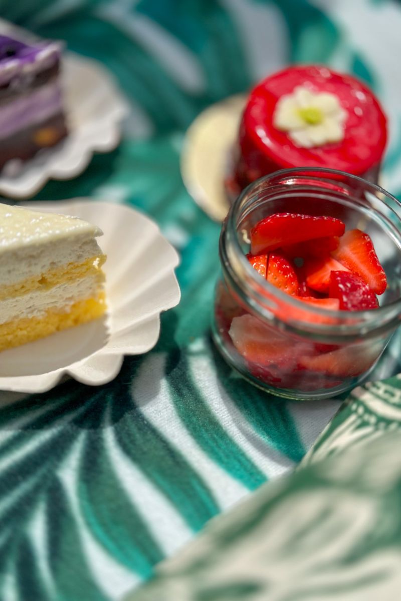 Sladki trenutki piknik paketa OBRAZI VRTNIC Od blizu fotografirane sladice na piknik odeji z zelenim listnim vzorcem. V ospredju je kozarček z narezanimi jagodami, ob njem rezina svetle torte, v ozadju pa rdeča sladica s cvetnim okrasjem in rezina vijolične torte.