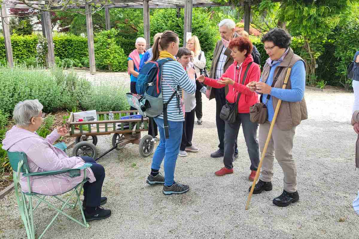 Čutno raziskovanje dišečega vrta Udeleženci v dišečem vrtu opazujejo in vonjajo rastline, ena udeleženka sedi ob vozičku z materiali.
