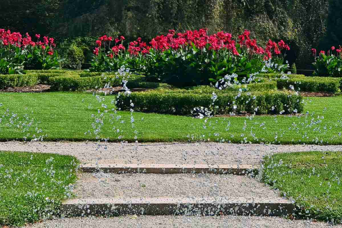 Arboretum Foto Mija Jerman-3 Vodni curki se dvigajo nad prodnato potjo v urejenem delu Arboretuma, v ozadju so zelene žive meje in rdeče cvetoče zasaditve.