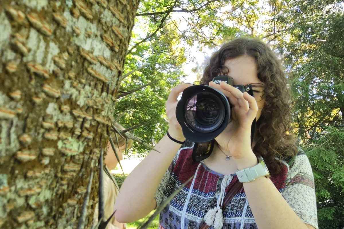 Udeleženka fotografske delavnice fotografira lubje drevesa od blizu, v ozadju so zelene krošnje Arboretuma Volčji Potok.