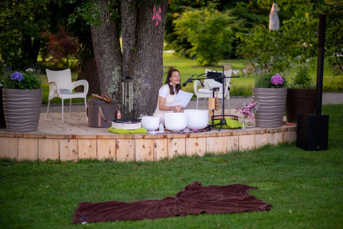 Ambientalna glasba med vrtnicami, foto Nika Halas Sandra Prešeren sedi na lesenem odru pod drevesi in izvaja ambientalno glasbo s holističnimi glasbili. Pred njo so postavljene zvočne posode, mikrofon in drugi instrumenti, na travi pred odrom pa leži odeja za poslušalce.