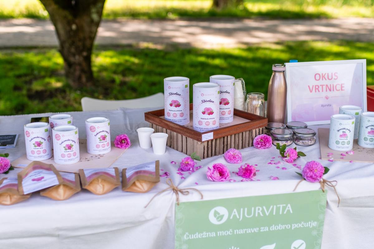 Okus vrtnice, Ajurvita foto Nika Halas Na stojnici Okus vrtnice so razstavljeni izdelki Ajurvita z vrtnicami, med njimi pločevinke, vrečke in kozarčki. Miza je okrašena z rožnatimi vrtnicami, cvetnimi listi in napisom »Okus vrtnice«.