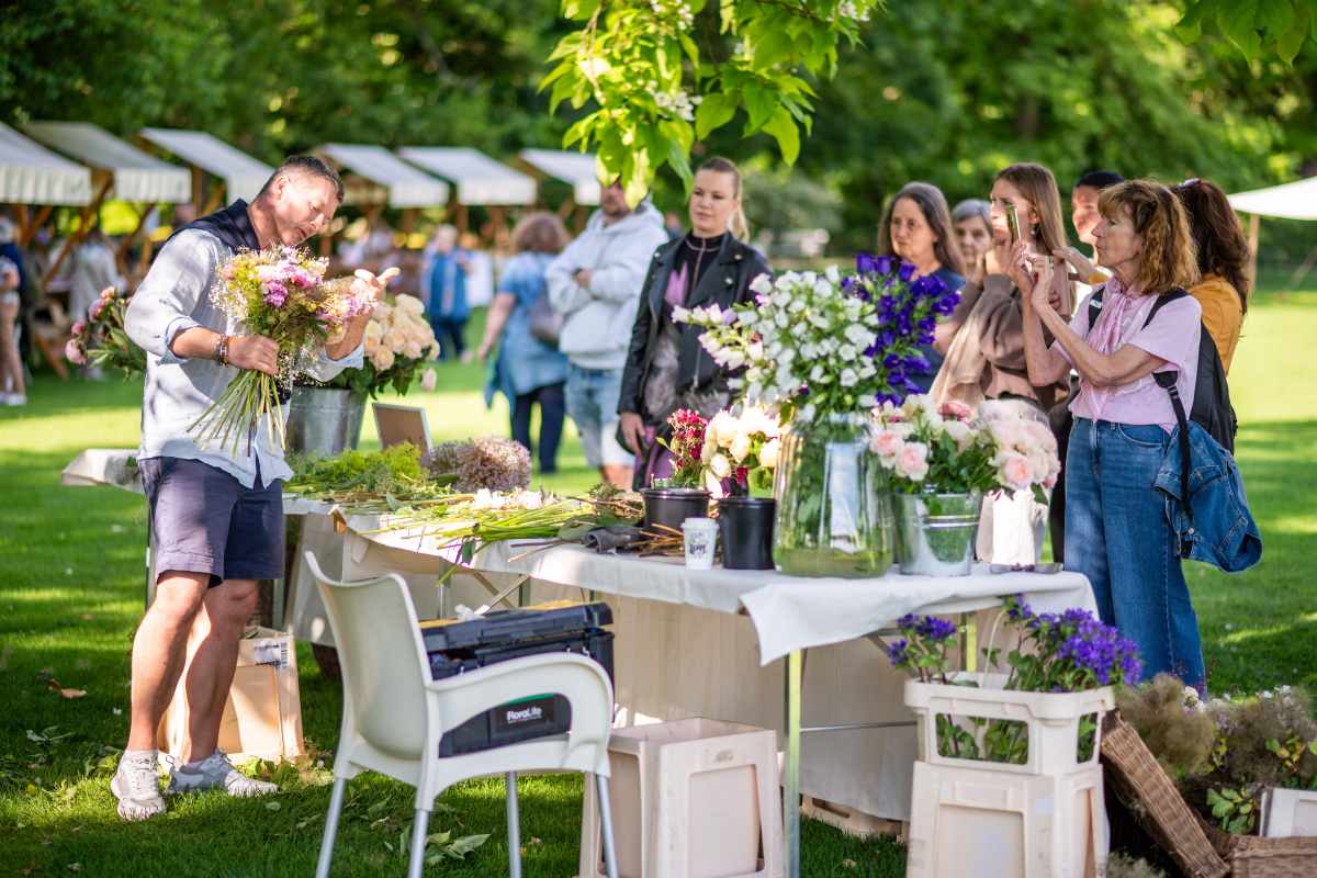 Florist na prostem med obiskovalci izdeluje šopek iz vrtnic in drugega cvetja. Na mizah so razporejene vrtnice, zelenje, cvetličarski pripomočki in pripravljeni cvetlični materiali, obiskovalci pa opazujejo prikaz izdelovanja šopkov.