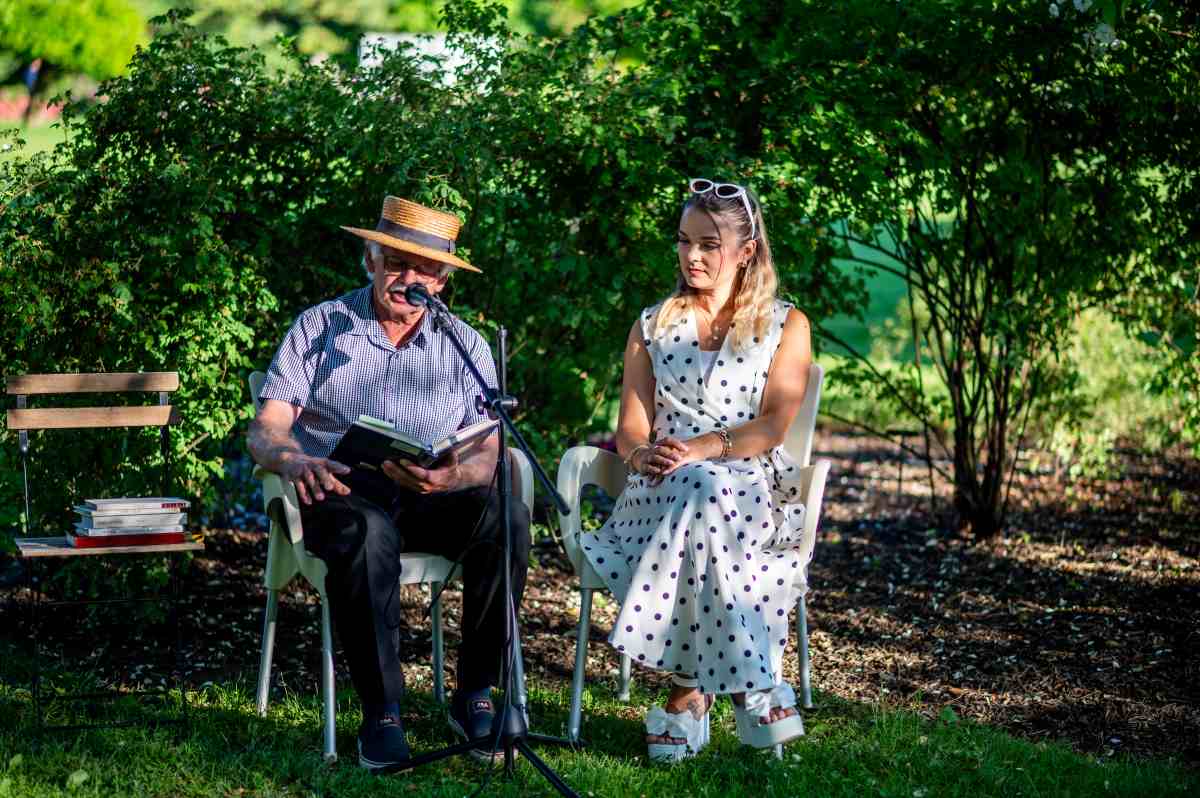 Zgodbe o vrtnicah, foto Nika Halas Moški s slamnikom sedi pod drevesi in ob mikrofonu bere iz knjige, ob njem sedi ženska v beli obleki s pikami. Ob strani je klop z zloženimi knjigami, prizor pa poteka v senci zelenja.