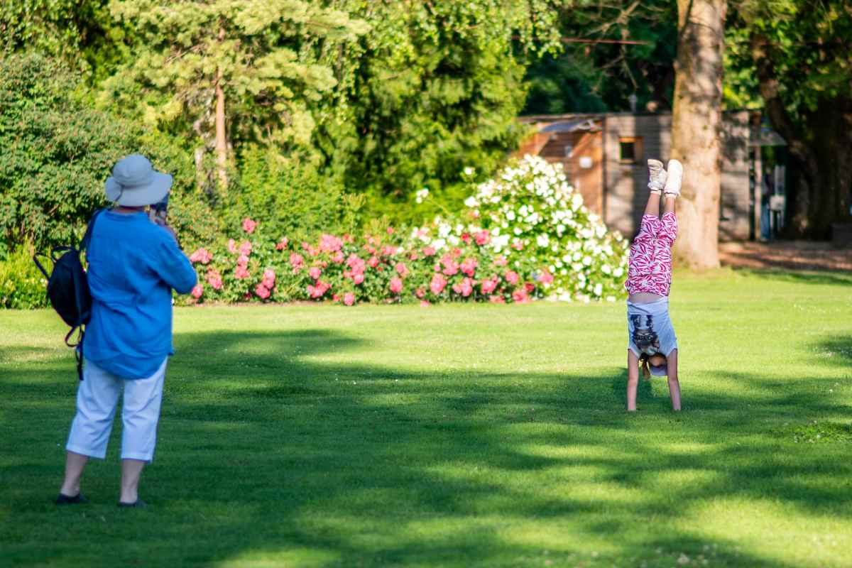 Igriv trenutek v Spodnjem rozariju, foto Nika Halas Otrok dela stojo na rokah na zeleni trati, v ozadju so cvetoče vrtnice, oseba v modri srajci pa ga fotografira.