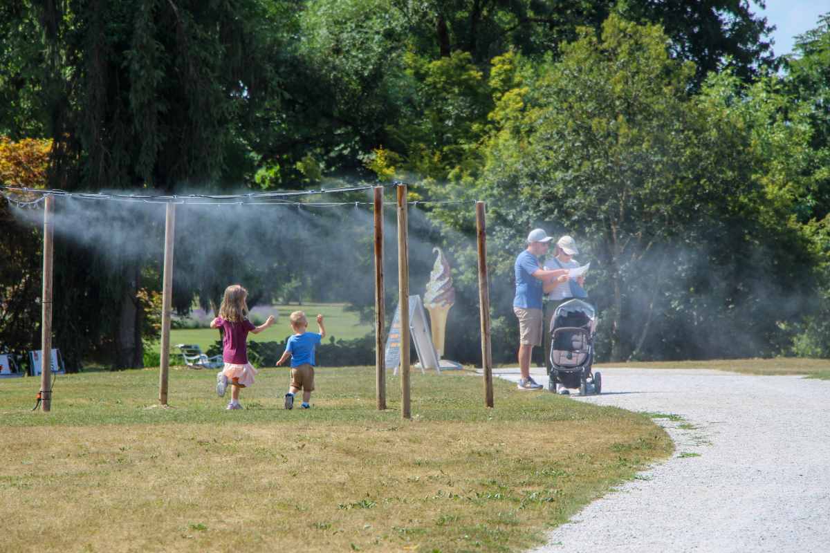 Družinski poletni izlet v Arboretumu Otroka tečeta skozi osvežilno vodno pršenje, v ozadju starša z vozičkom gledata zemljevid parka.