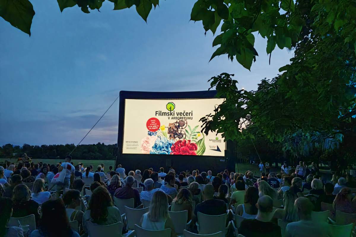 Filmski večer pod odprtim nebom Obiskovalci sedijo pred velikim filmskim platnom na prostem, obdani z drevesi in večernim nebom.