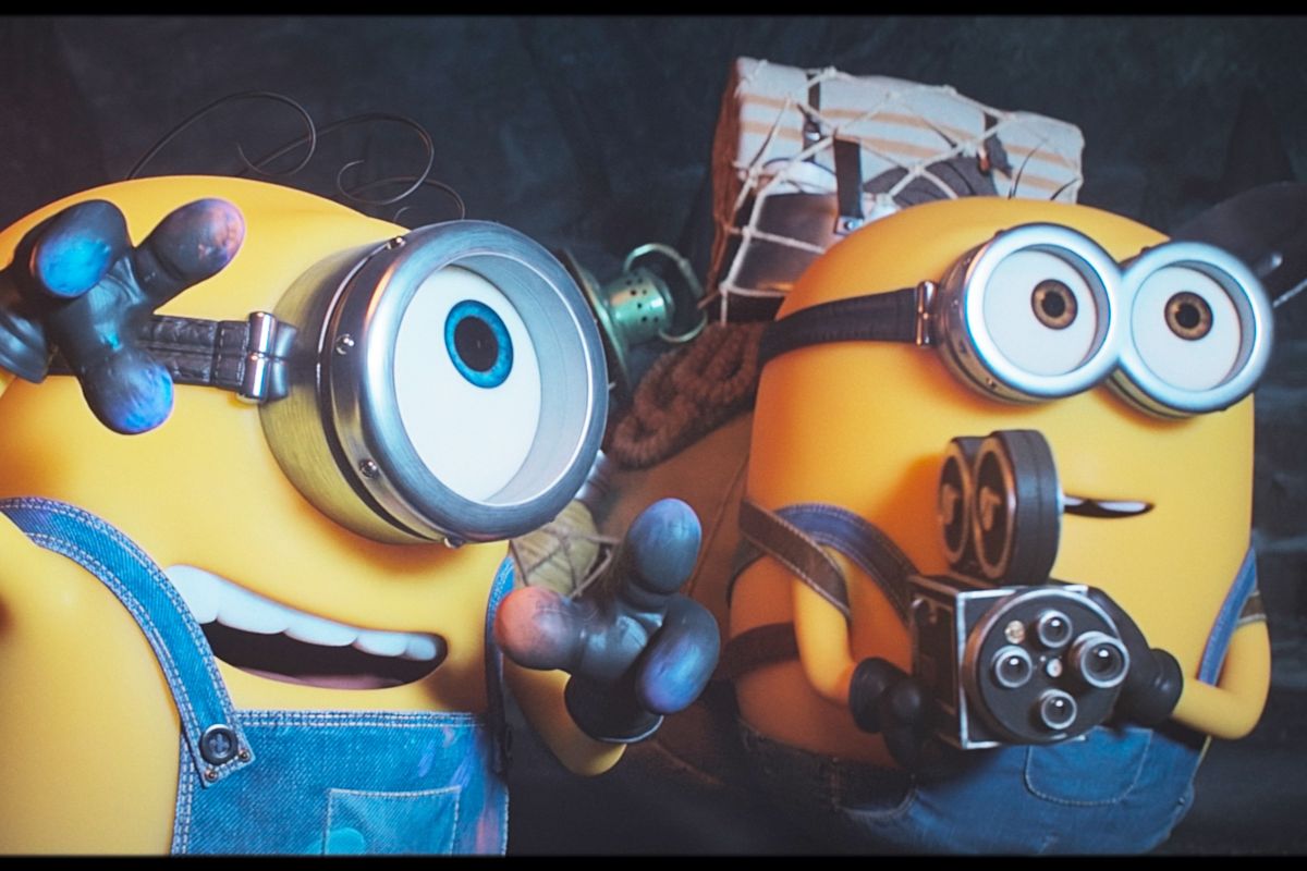 Prizor iz animiranega filma Minioni in pošasti prikazuje dva rumena Miniona z zaščitnimi očali. Eden maha proti gledalcu, drugi pa v rokah drži majhno kamero.