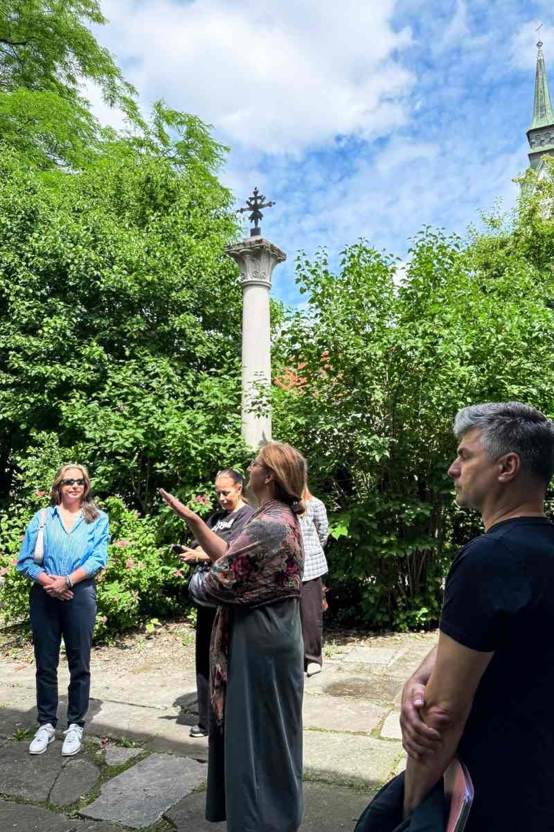 Plečnikov vrt Strokovnjakinja govori skupini obiskovalcev na vrtni terasi ob kamnitem stebru, drevesih in grmovnicah.