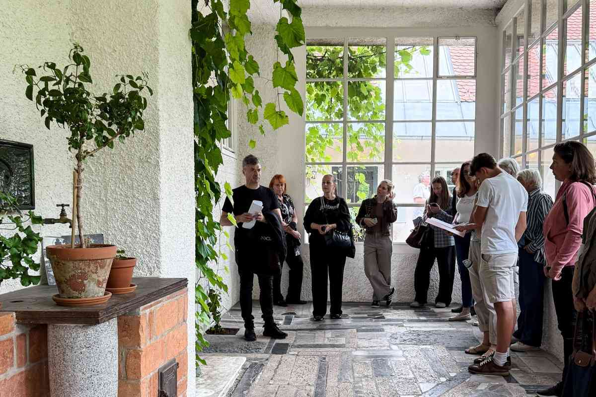 Vodenje v zastekljenem delu Plečnikove hiše Skupina obiskovalcev posluša razlago v svetlem zastekljenem prostoru Plečnikove hiše, obdanem z lončnicami in vzpenjavkami.
