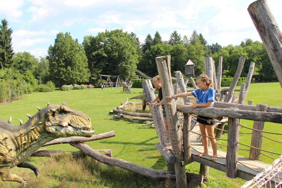 Dinozavri v Arboretumu Otroka stojita na lesenem igralu in opazujeta velikega modela dinozavra na zelenem travniku v Arboretumu.