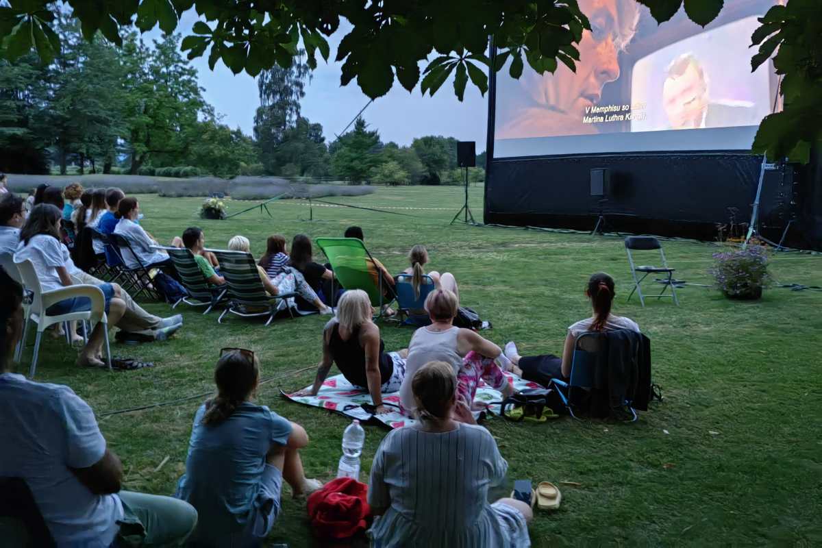 Film med drevesi Obiskovalci sedijo na odejah in stolih pred velikim filmskim platnom, prizorišče obdajajo drevesa in večerna svetloba.